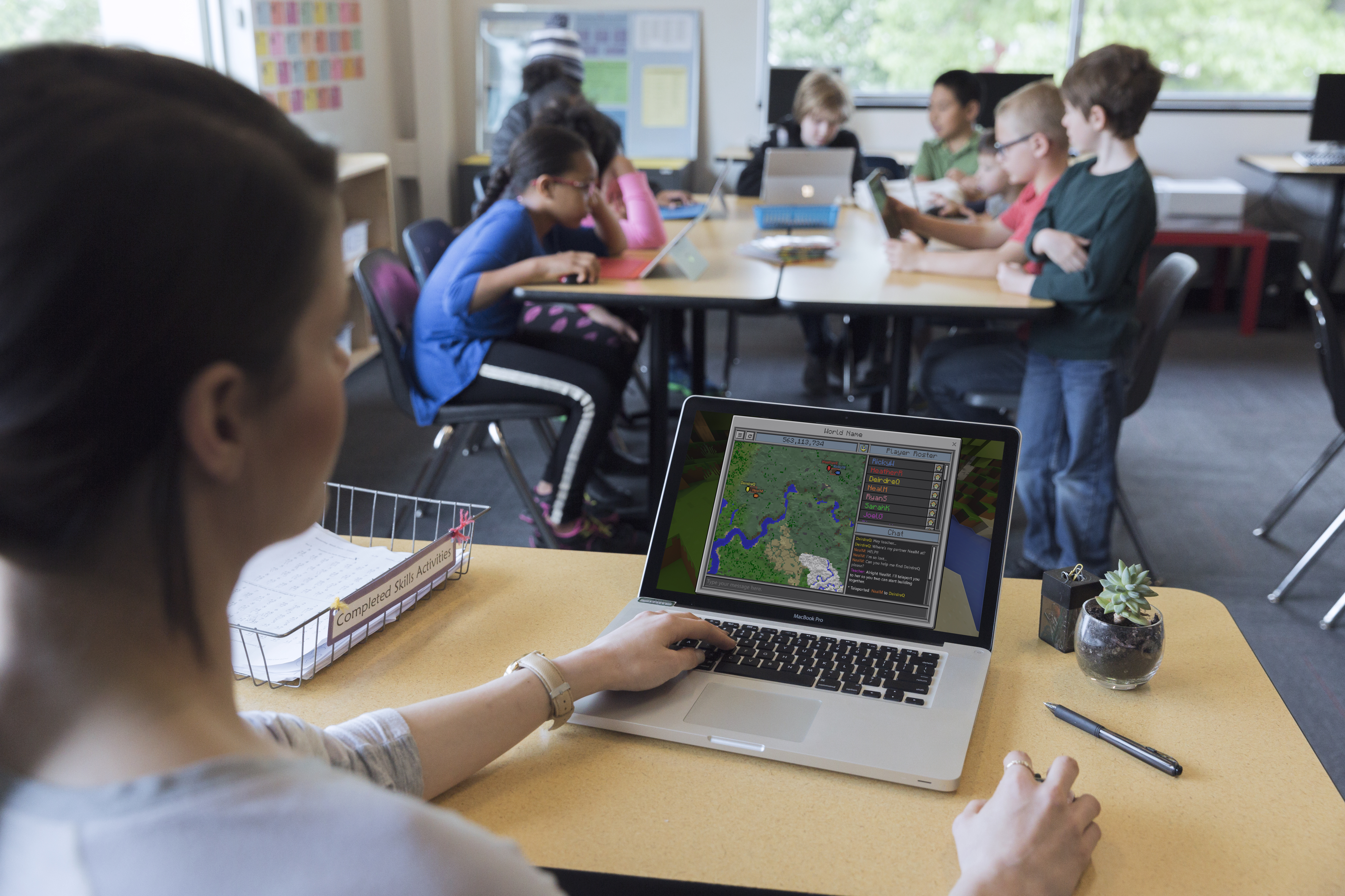 Teacher sitting at a desk with Classroom Mode for Minecraft open and students in the background sitting at their desks with Minecraft Education open.
